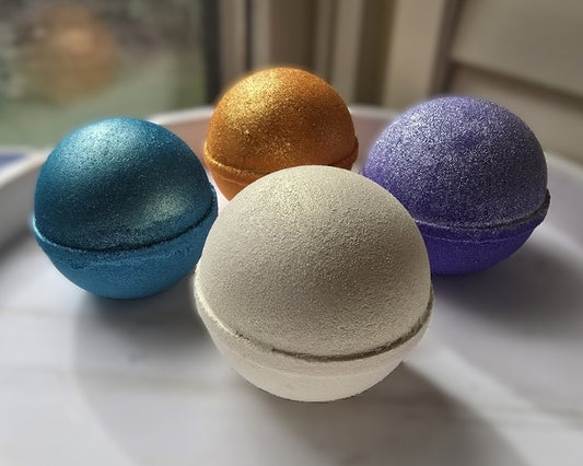 Four glittery bath bombs in blue, gold, white, and purple on a light surface.