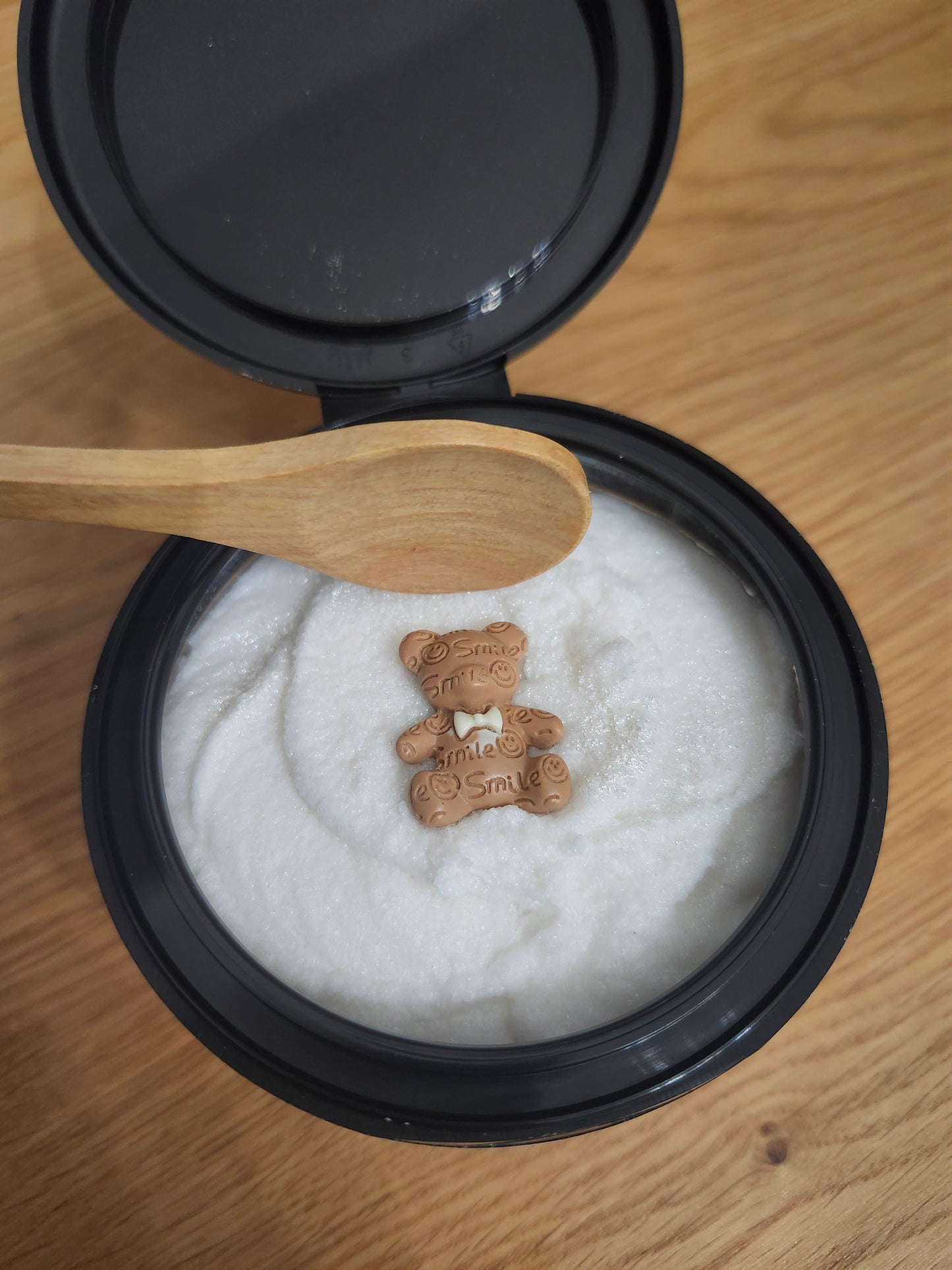 Container of white sugar scrub with a wooden spoon and a small brown bear-shaped charm on a wooden surface