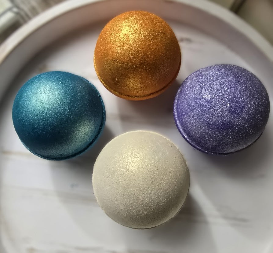 Four glittery bath bombs in blue, gold, white, and purple on a white plate.