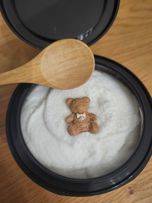 Container of white sugar scrub with a small charm on top, wooden spoon beside it on a wooden surface.